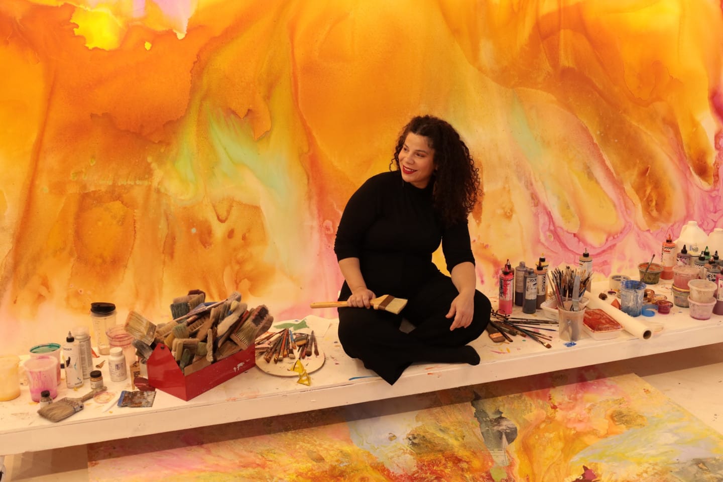 Artist Firelei Baez sits in a studio amongst paints, in front of a bright yellow painting