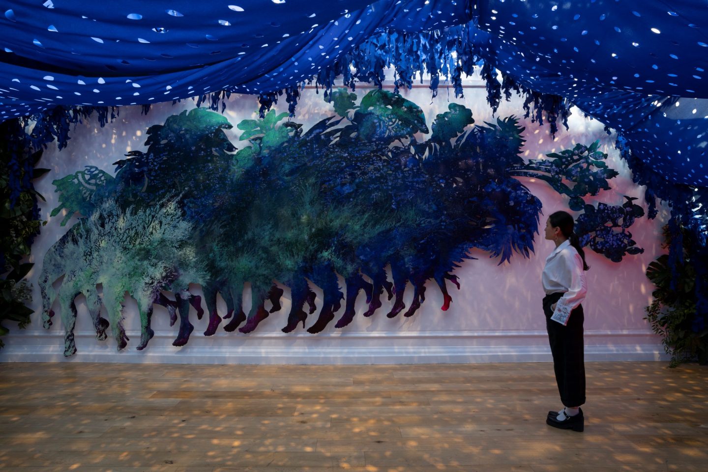 A large art gallery space with an immersive installation. The art work has blue tarp hanging from the ceiling. One person looking at an aluminium painted mythical creature.